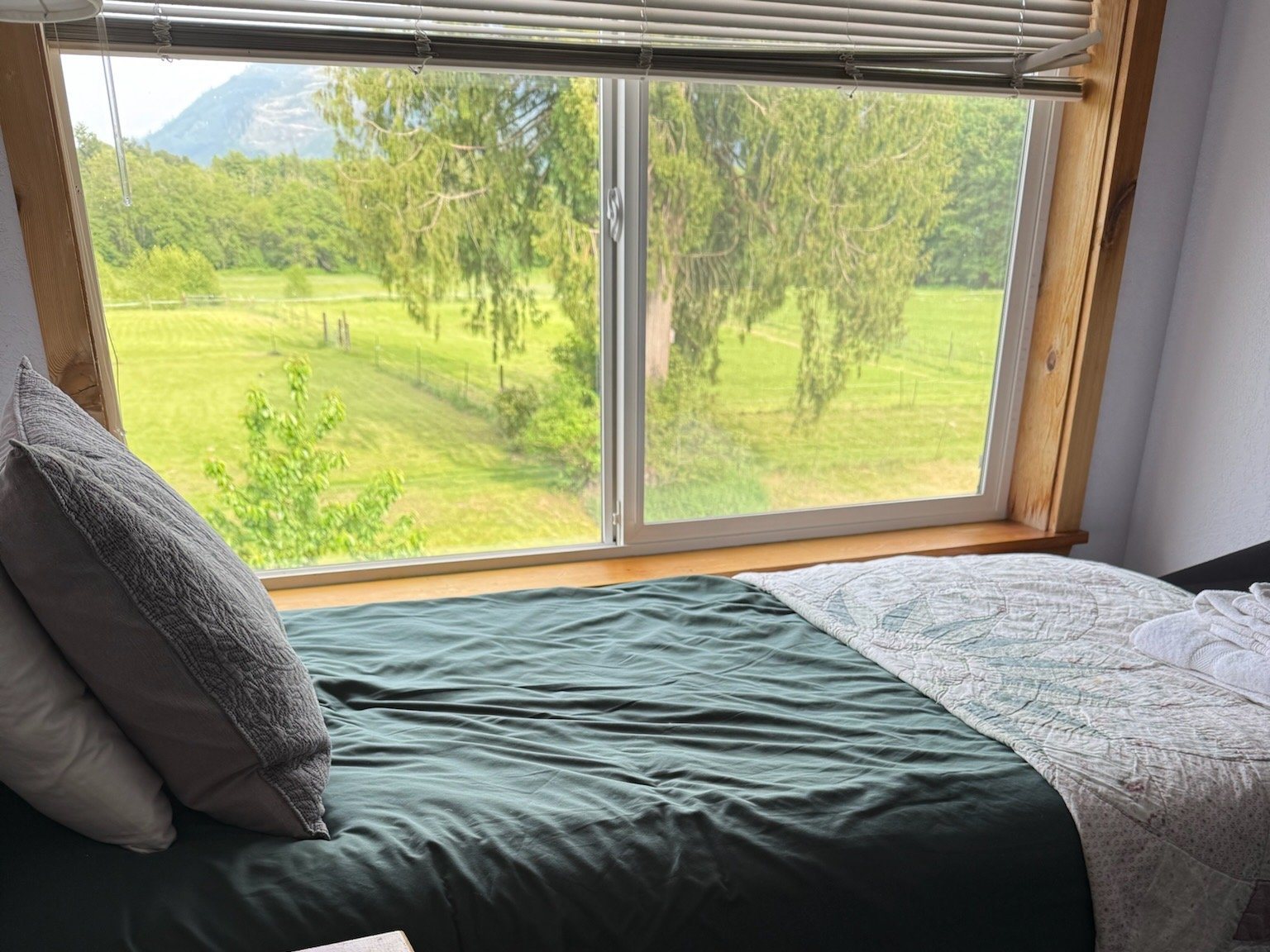 Morning view from an upstairs bedroom: a bed below a wide window looking out over green pasture, tall trees, and a mountain peak